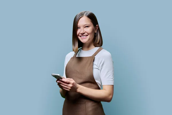 Portrait of young woman in apron holding smartphone in hands on blue background. Smiling female using mobile phone, texting receiving sending order. Technologies applications service, small business
