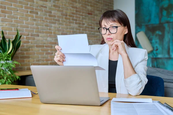 Serious mature businesswoman reading important official letter sitting at desk in office. Business work banking services taxes, contract statement agreement investment deal, paperwork correspondence