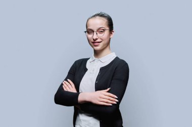 Gri arka planda gözlüklü, kendine güvenen gülümseyen genç kız portresi. Pozitif kadın, 16-17 yaşlarında çapraz kollu lise öğrencisi bir gençle kameraya bakıyor.