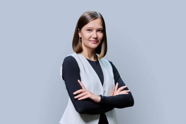 Kollarını kavuşturup gri stüdyo arka planındaki kameraya bakan kendine güvenen, güler yüzlü genç bir kadın. Güzellik, iş, iş... Girişimcilik... İşletme uzmanlığı...