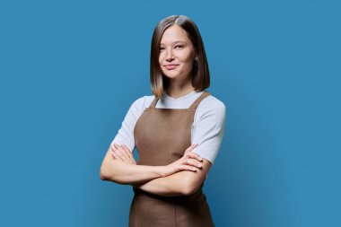 Önlüklü genç bir kadın mavi stüdyo arka planındaki kameraya bakıyor. Kendine güvenen küçük işletme sahibi, kolları bağlı şekilde poz veren bir servis çalışanı. İş, iş, meslek, personel kavramı