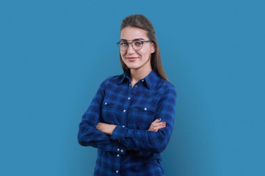 Mavi stüdyo arka planında gülümseyen pozitif kadının portresi. Kendine güveni tam bir kadın, kolları çapraz gömlekli, rahat bir şekilde kameraya bakıyor. Gençlik, yaşam tarzı, 30 yaşındaki insanlar konsepti.