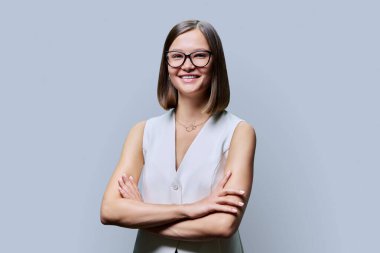 Gözlüklü, kollarını kavuşturup gri stüdyo arkaplanındaki kameraya bakan, kendine güvenen, modaya uygun gülümseyen genç bir kadın. Güzellik, iş, iş... Girişimcilik... İşletme uzmanlığı...