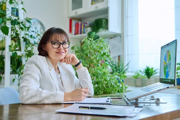 Portrait of mature businesswoman working at home on computer laptop. Smiling middle-aged female looking at camera sitting at desk in home office. Remote business work, career, management, marketing