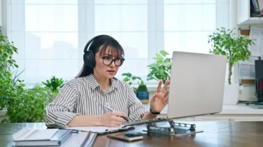 Merkez ofisteki bilgisayarda çalışan orta yaşlı kulaklıklı bir kadın web kamerasına bakarak konuşuyor. Çevrimiçi toplantı, uzak çalışma finansal danışmanlık danışmanlık blog yazarlığı, zihinsel terapi