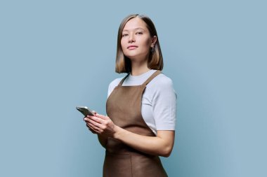 Mavi arka planda akıllı telefonu elinde tutan önlüklü genç bir kadının portresi. Ciddi bir kadın cep telefonu kullanıyor, kameraya bakıyor. Teknolojiler, uygulamalar, hizmet, küçük işletmeler, iş