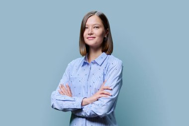 Kollarını kavuşturup mavi stüdyo arka planındaki kameraya bakan kendine güvenen genç bir kadının portresi. Yirmili yaşlardaki insanlar, gençler, iş, eğitim, moda, yaşam tarzı kavramı