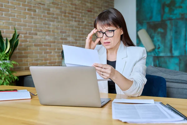 Serious mature businesswoman reading important official letter sitting at desk in office. Business work banking services taxes, contract statement agreement investment deal, paperwork correspondence