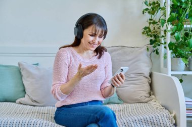 Kulaklıklı orta yaşlı bir kadın görüntülü konuşma konferansı için akıllı telefon kullanıyor. Yetişkin bir kadın telefon ekranında konuşuyor. Vlog, çevrimiçi iletişim, modern iletişim teknolojileri kavramı