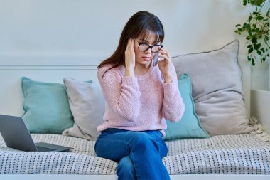 Ciddi bir yetişkin kadın telefonda konuşuyor, evdeki kanepede oturuyor. Ciddi konuşmalar, sorunlar, zorluklar, çatışmalar, kayıplar