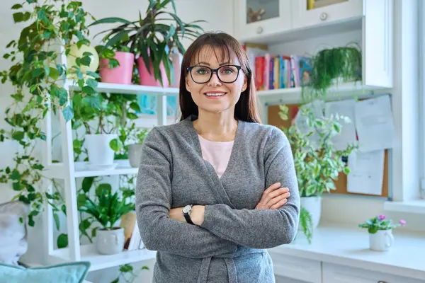 Portrait of confident smiling middle-aged woman in glasses, casual clothes with crossed arms, looking at camera in home interior. 40s age, lifestyle, health, mature lady concept