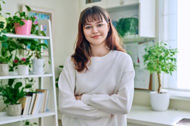 18-20 yaşlarındaki genç bir kızın portresi. Evde kollarını kavuşturmuş kameraya bakıyor. Burun piercingli güzel bir kız. Makyajlı saç stili. Gençlik yaşam tarzı güzellik modası