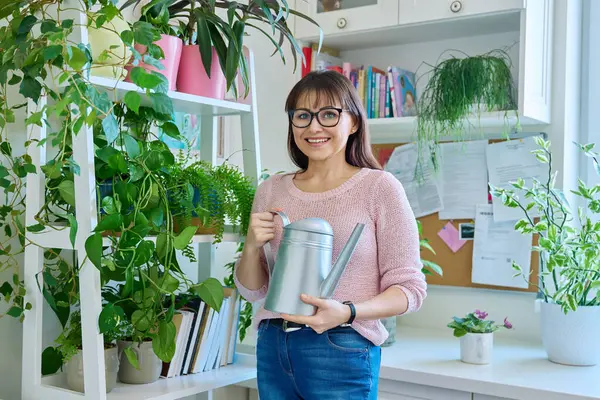 Middle-aged woman watering pots with plants from watering can, home interior. Smiling female looking at camera. Home landscaping, eco green trends, lifestyle concept