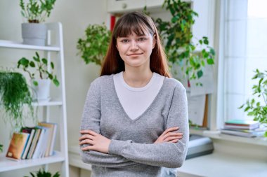 18-20 yaşlarındaki genç bir kızın portresi. Evde kollarını kavuşturmuş kameraya bakıyor. Burun piercingli güzel bir kız. Makyajlı saç stili. Gençlik yaşam tarzı güzellik modası