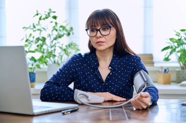 Tansiyonunu ölçen orta yaşlı bir kadın, iş yerinde bilgisayarla masada oturuyor. Sağlık, bakım, olgun insanlar konsepti