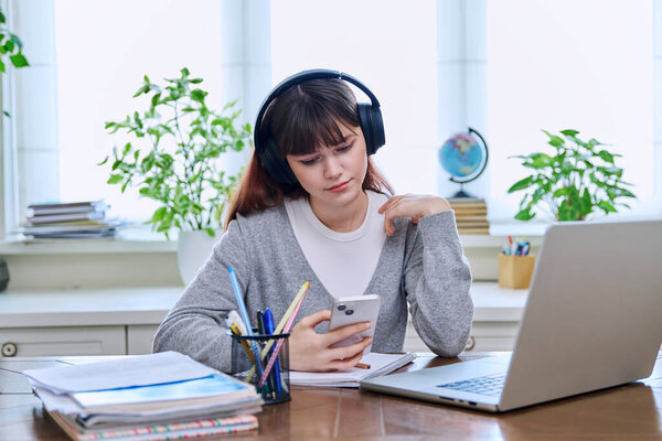 Teenage college student girl wearing headphones with smartphone in hands sitting at desk with computer with textbooks notebooks. Technology, education, training, e-learning, youth concept