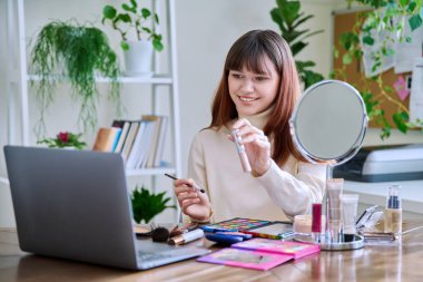 Dizüstü bilgisayara kozmetik gösteren genç bir bayan, evde masada oturuyor. Güzellik blogu vlog, çevrimiçi kurs, dekoratif kozmetik ürünlerinin profesyonel kullanımı üzerine webinar eğitimi