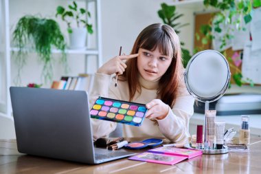 Dizüstü bilgisayara kozmetik gösteren genç bir bayan, evde masada oturuyor. Güzellik blogu vlog, çevrimiçi kurs, dekoratif kozmetik ürünlerinin profesyonel kullanımı üzerine webinar eğitimi