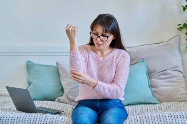 Orta yaşlı bir kadın dirsek eklemine dokunuyor, evdeki kanepede oturuyor. Ağrı sendromları: artrit, osteoartrit, romatizma, çürük. Sağlık, tedavi, bakım, olgun insanlar konsepti