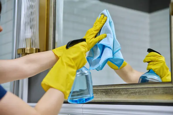 Close-up hands in protective gloves cleaning bathroom, washing mirror with spray bottle and professional rag for cleaning glass without streaks. Housekeeping housework housecleaning service concept