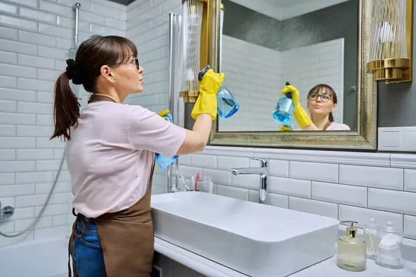 中年妇女穿着围裙 手套清洁浴室 用专业的抹布洗涤剂清洗镜子 女家庭妇女清扫房屋 工作场所的服务人员 料理家务 — 图库照片