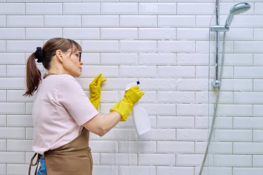 Önlüklü orta yaşlı bir kadın, banyoyu profesyonel deterjanla temizleyen eldivenler duşta fayansları temizliyor. Kadın ev hanımı temizlikçi, iş yerinde servis işçisi. Oda servisi ev işi