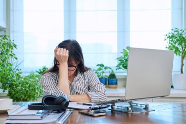 İş yerinde bilgisayarlı dizüstü bilgisayarla el ele tutuşan üzgün, olgun bir kadın. Orta yaşlı insanlar, stres sıkıntısı, baş ağrısı sağlık, yaşam tarzı konsepti