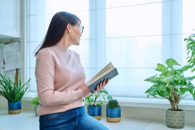 Orta yaşlı bir kadın evinde, pencerenin yanında kurgu kitabı okuyor. Düşünceli bir şekilde pencereden dışarı bakıyor. Kış mevsimi.