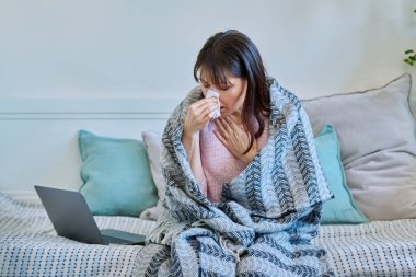 Hasta, olgun bir kadın. Burnunu sümkürerek hapşırıyor. Evdeki kanepede oturuyor. Soğuk algınlığı, sonbahar kışı mevsimsel solunum hastalıkları, tedavi ve bakım