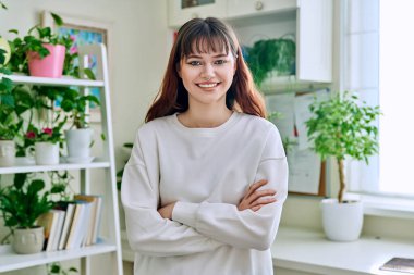 18-20 yaşlarındaki genç bir kızın portresi. Evde kollarını kavuşturmuş kameraya bakıyor. Burun piercingli güzel bir kız. Makyajlı saç stili. Gençlik yaşam tarzı güzellik modası