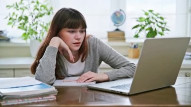 Sinirli genç kız öğrenci, açık bir dizüstü bilgisayarın yanında masada. Genç bir kız bilgisayar ekranına bakıyor, haber bekliyor.