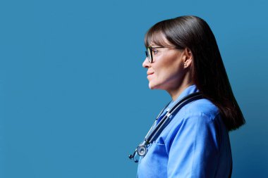 Gülümseyen orta yaşlı, kendine güvenen kadın hemşirenin portresi mavi stüdyo arka planında, fotokopi alanında. Tıbbi hizmetler, sağlık, mesleki yardım, tıbbi bakım kavramı