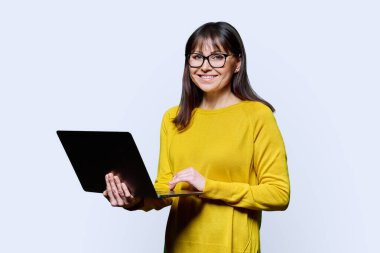 Beyaz stüdyo arka planında dizüstü bilgisayar kullanan sarı elbiseli orta yaşlı bir kadın. Kameraya bakıp gülümseyen olgun bir kadın. İletişim için teknoloji ağı iş hayatı tarzı 40 'lı yılların insan kavramı