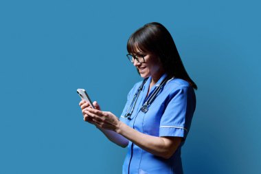Gülümseyen kadın hemşire akıllı telefon kullanıyor, profil görüntüsü mavi stüdyo arka planı, fotokopi odası. Mobil uygulamalar, tıbbi hizmetlerdeki teknolojiler, sağlık, profesyonel yardım, tıbbi bakım