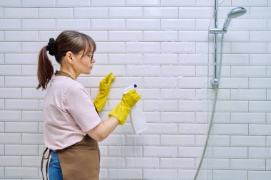 Önlüklü orta yaşlı bir kadın, banyoyu profesyonel deterjanla temizleyen eldivenler duşta fayansları temizliyor. Kadın ev hanımı temizlikçi, iş yerinde servis işçisi. Oda servisi ev işi