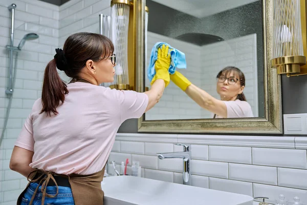 中年妇女穿着围裙 手套清洁浴室 用专业的抹布洗涤剂清洗镜子 女家庭妇女清扫房屋 工作场所的服务人员 料理家务 — 图库照片