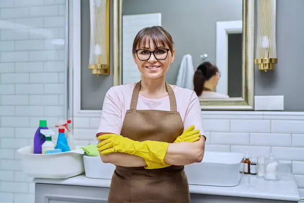 Middle-aged woman in gloves apron with crossed arms in bathroom. Female preparing for routine house cleaning, housecleaning service worker posing at workplace. Housekeeping housework cleaning