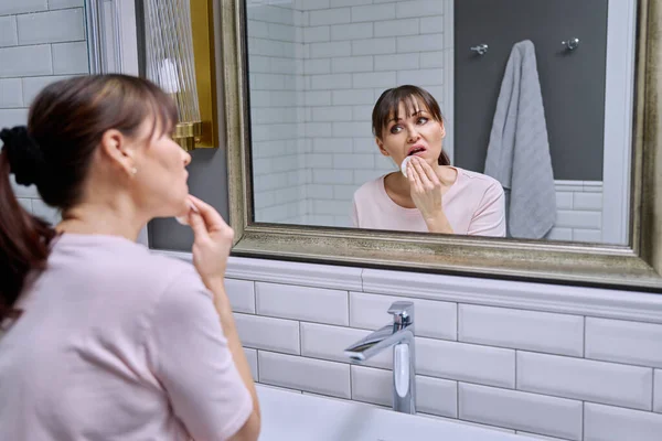 Middle-aged woman with cotton pad cleans her face, lips from lipstick, looking in the mirror at home in bathroom. Mature age, beauty routines, makeup removal concept