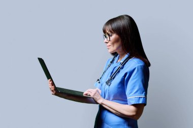 Gülümseyen kadın hemşire dizüstü bilgisayar kullanıyor, profil görüntüsü gri stüdyo arka planında. Başvurular, tıbbi hizmetlerdeki teknolojiler, sağlık, mesleki yardım, tıbbi bakım kavramı