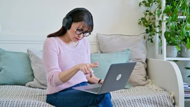 中年妇女坐在家里的沙发上 头戴耳机 在笔记本电脑上举行视频电话会议 Vlog Work Leisure Online Communication Modern Technologies — 图库视频影像