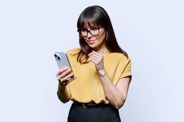 Beyaz arka planda akıllı telefon kullanan orta yaşlı ciddi bir kadın. Yetişkin bir kadın elinde telefona bakıyor. Teknolojiler, mobil uygulamalar, uygulamalar, internet işi, iş fırsatı iletişimi.