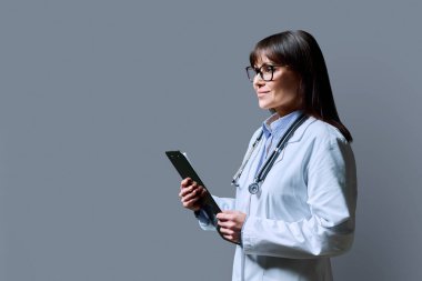 Beyaz laboratuvar önlüklü gülümseyen, kendine güvenen kadın doktorun profil portresi. Elinde gri stüdyo arka planında bir pano var. Kopya alanı. Sağlık, ilaç, personel, tedavi, tıbbi hizmetler kavramı