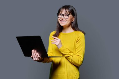 Sarı elbiseli orta yaşlı kadın gri stüdyo arka planında dizüstü bilgisayar kullanıyor. Kameraya bakıp gülümseyen olgun bir kadın. İletişim için teknoloji ağı iş hayatı tarzı 40 'lı yılların insan kavramı