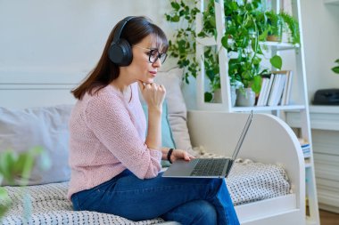 Kulaklıklı, orta yaşlı, ciddi bir kadın bilgisayarında görüntülü konuşma konferansı yapıyor. Evdeki koltukta oturuyor. Vlog, iş, eğlence, çevrimiçi iletişim, modern teknoloji kavramı