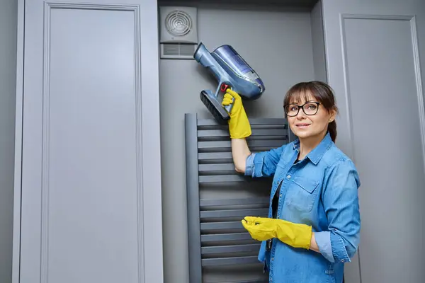 Woman cleaning wall-mounted dusty hood ventilation grill in bathroom with vacuum cleaner. Housekeeping, housework, housecleaning, cleaning service concept
