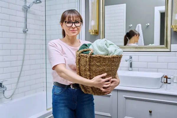 Middle aged woman with basket of towels for washing at home in bathroom, smiling female looking at camera. Housekeeping, laundry, domestic life, lifestyle concept