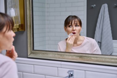 Pamuklu ped kullanan orta yaşlı bir kadın yüzünü temizliyor, rujdan dudakları dökülüyor, banyodaki aynaya bakıyor. Olgun yaş, güzellik rutinleri, makyaj temizleme konsepti