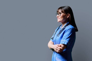Kollarını kavuşturmuş gülümseyen orta yaşlı, kendine güvenen kadın hemşirenin portresi gri stüdyo arka planında, fotokopi alanında. Sağlık hizmetleri profesyonel yardım tıbbi bakım kavramı
