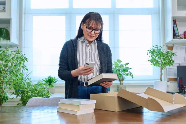 Middle aged woman with smartphone unpacking box with books, purchase from bookstore, online shopping, in home interior. Unpacking, postal delivery, online shopping, books literature concept