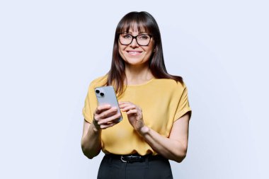 Beyaz arka planda akıllı telefon kullanan orta yaşlı gülümseyen bir kadın. Yetişkin bir kadın elinde telefonla kameraya bakıyor. Teknolojiler mobil uygulamalar İnternet işi eğlence iletişimi